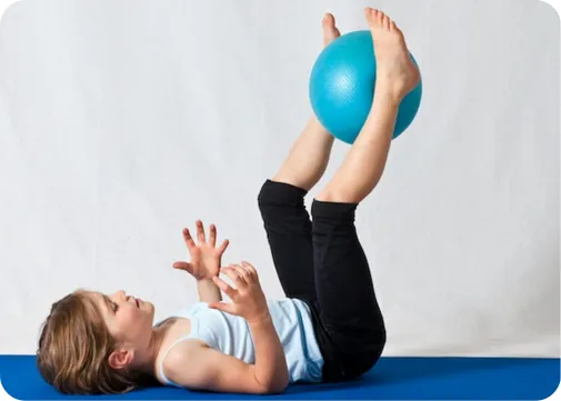 Child lying on her back, holding a therapy ball between her legs during a physiotherapy exercise